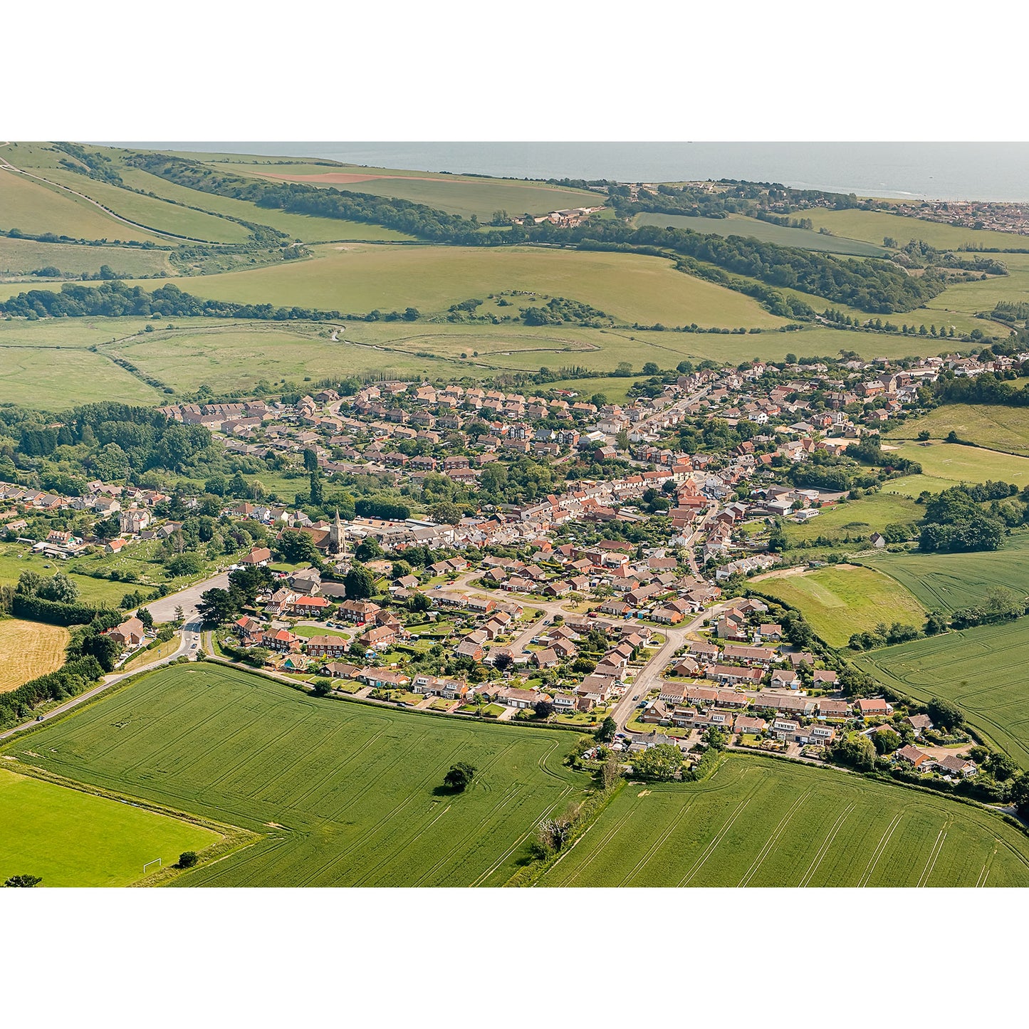 The product "Brading" by Available Light Photography captures an aerial view of a small Isle of Wight town, showcasing green fields and rolling hills lightly veiled by morning mist under a clear sky.