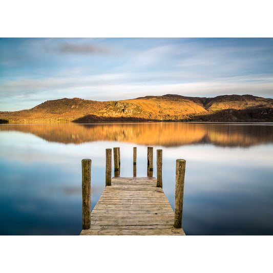 A serene view of a wooden jetty extending into Derwent Water on the Isle of Wight with reflections of surrounding hills under a clear sky by Available Light Photography.