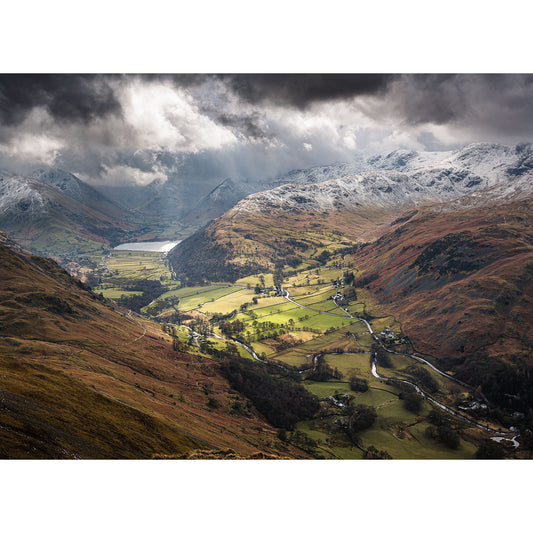 A scenic view of a valley with snow-capped mountains, a patchwork of fields, and a winding river under a dramatic cloudy sky captured by Available Light Photography's Patterdale.