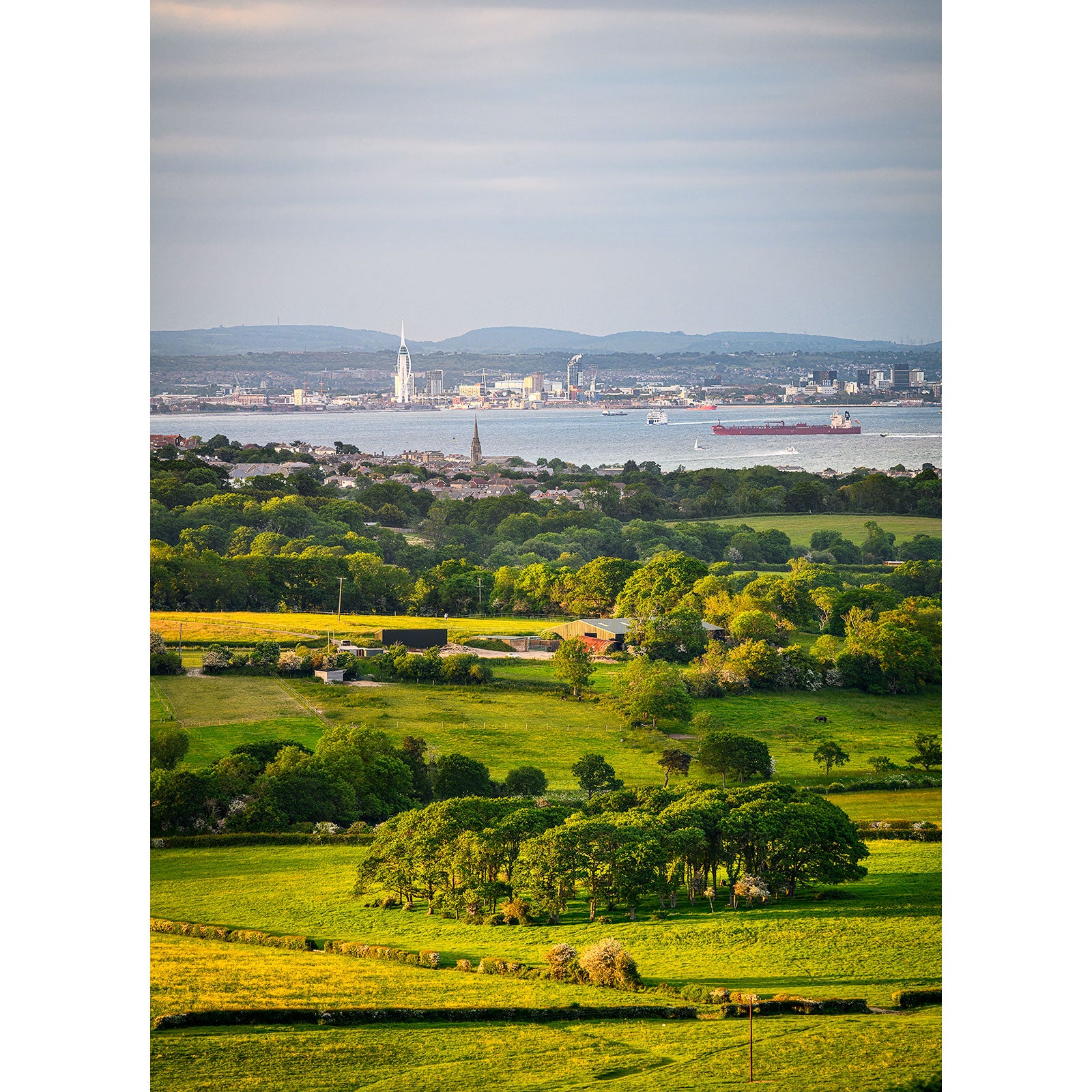 Lush green foreground with scattered trees leading to a cityscape and industrial harbor in the distance, under a cloudy sky on the Isle of Wight. - View from Ashey Down by Available Light Photography