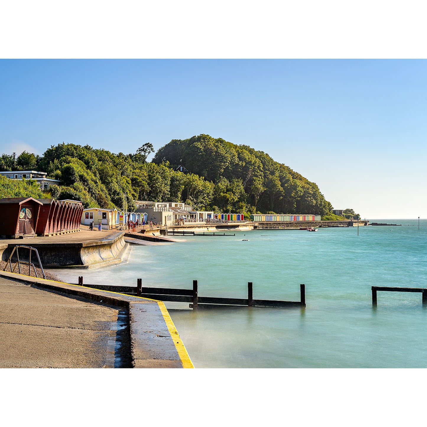 Coastal scene showing a calm blue sea, a concrete pier, colorful beach huts, and a blue sky on a sunny day at Colwell Bay by Available Light Photography on the Isle of Wight.