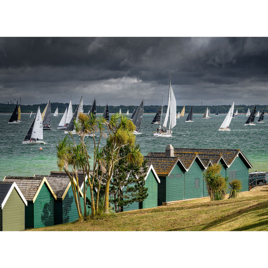 Sailboats racing in the Round the Island Race near the Isle of Wight, under a stormy sky, with colorful beach huts in the foreground. Captured by Available Light Photography.