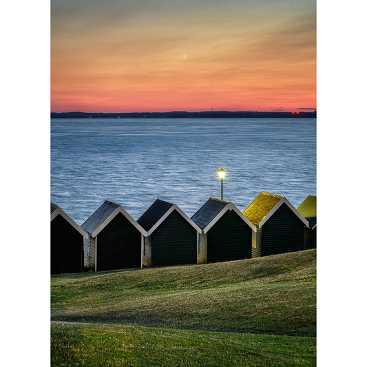 Row of colorful beach huts overlooking a calm sea at sunset with a crescent moon in the sky on Gurnard by Available Light Photography.