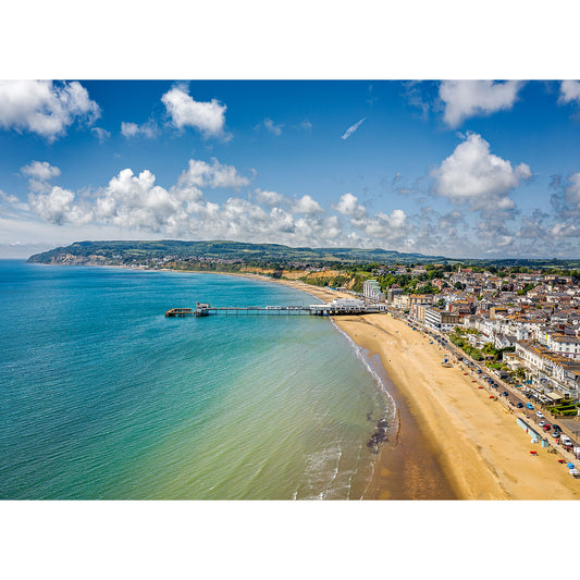 Aerial view of Sandown, a coastal town with a pier extending into the sea, sandy beachfront, and a backdrop of hills under a partly cloudy sky on Wight by Available Light Photography.