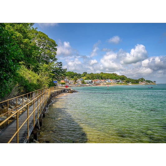 A tranquil coastal scene of Seagrove Bay on the Isle of Wight, with a wooden railing leading along the shoreline towards a small village with boats moored in the clear water. (Available Light Photography)