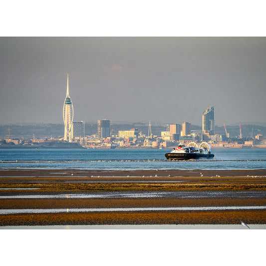 An Incoming Hovercraft, Ryde travels across a beach with a city skyline, featuring a tall, spire-like building, in the background. In the distance, the silhouette of Gascoigne Isle emerges. Crafted by Available Light Photography.
