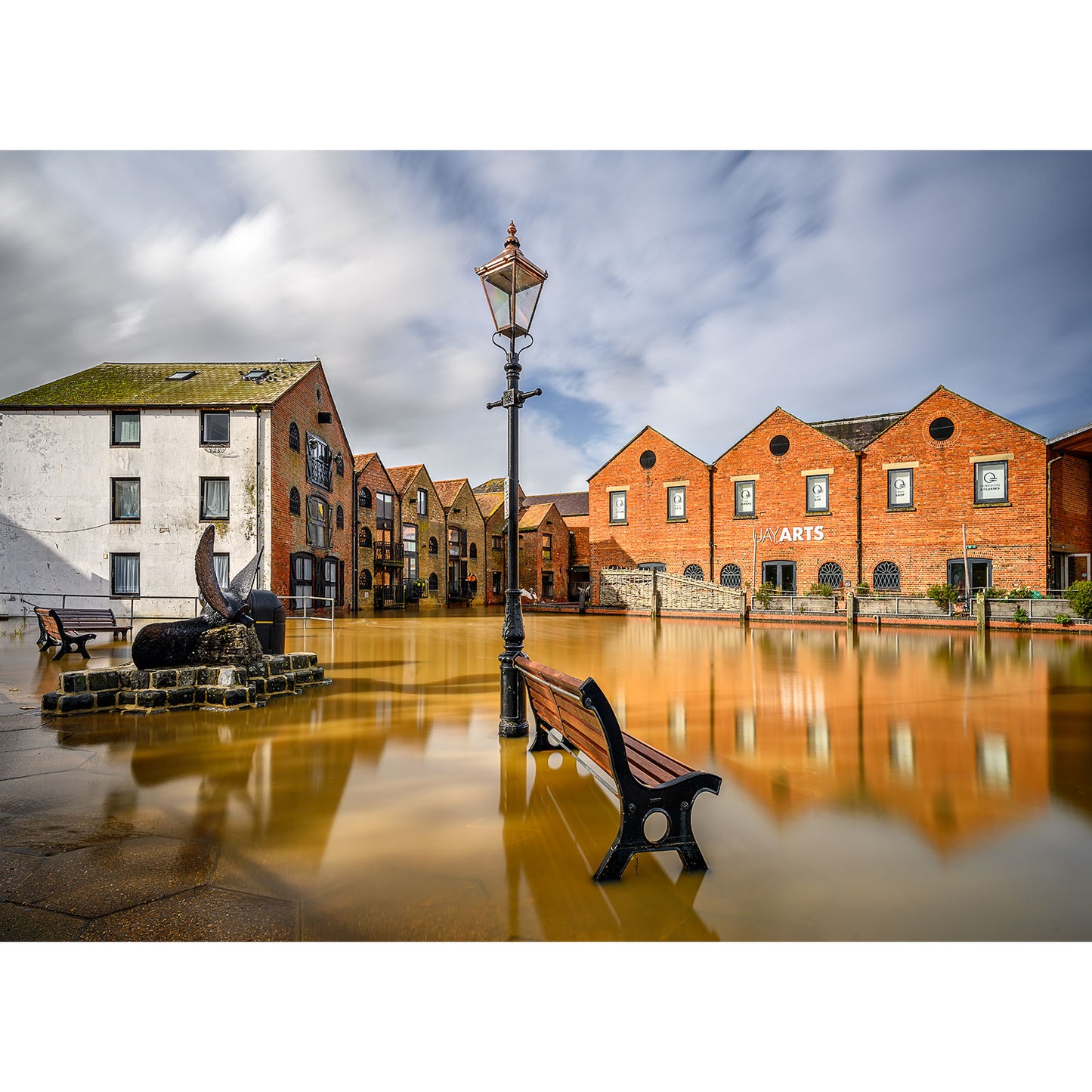 Flooded urban area with buildings, a lamppost, and a bench partially submerged in water at Newport Quay on the Isle of Wight by Available Light Photography.