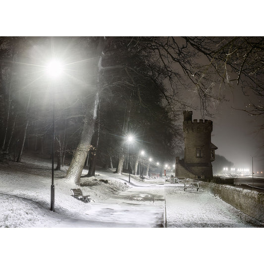 A wintry night scene with snow-covered ground, bare trees, illuminated by streetlights, and an Appley Tower in the background by Available Light Photography.
