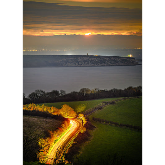 A winding road illuminated by car headlights cuts through a rural landscape on the Isle of Wight at dusk, with a sunset over distant hills and a calm sea, featuring "Moonrise over Culver Cliff" by Available Light Photography.