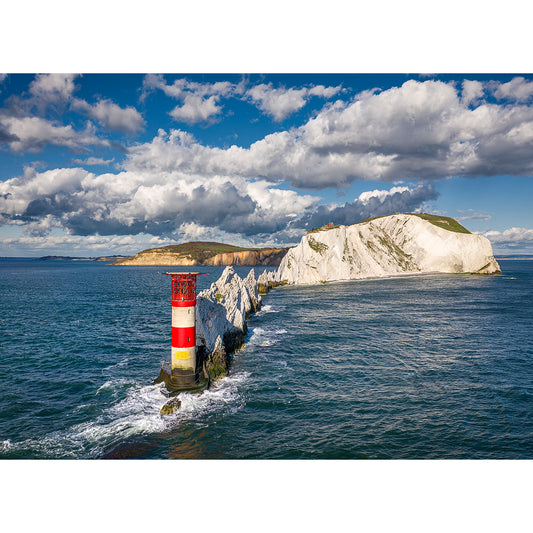A red and white lighthouse stands on a rocky cliff at the end of a jagged shoreline, with a backdrop of sunlit white cliffs under a cloudy sky captured by Available Light Photography's "The Needles.