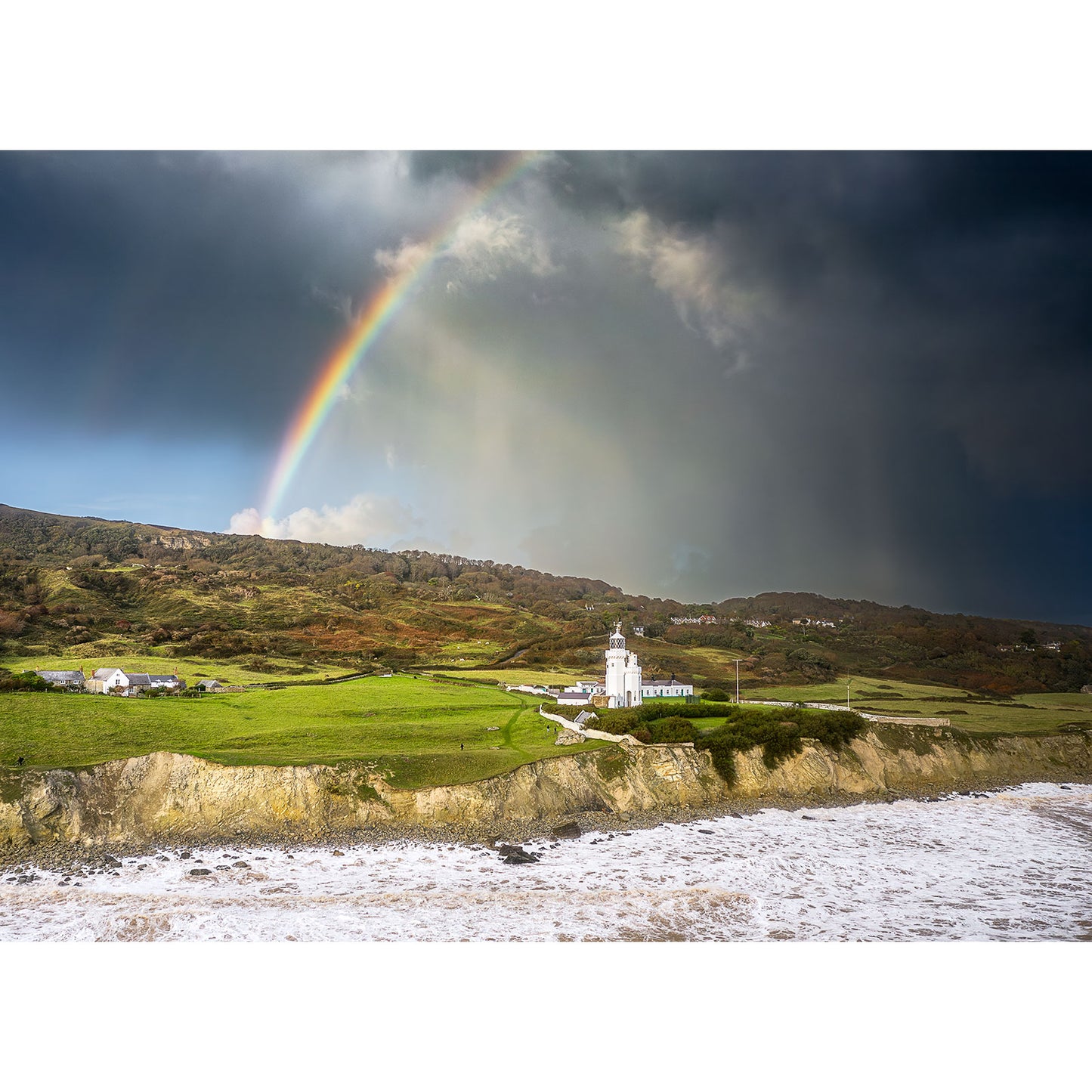 A rainbow arches over a coastal scene featuring St. Catherine's Lighthouse, green fields, and crashing waves under a dramatic sky—captured by Available Light Photography. No SEO keywords were found in the description provided.