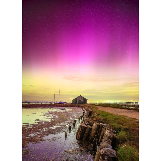 A wooden building stands near the water under a sky illuminated by pink and purple auroras, with a pathway leading to it and silhouetted boats in the background. This enchanting scene is captured perfectly in The Northern Lights, Newtown Creek by Available Light Photography.