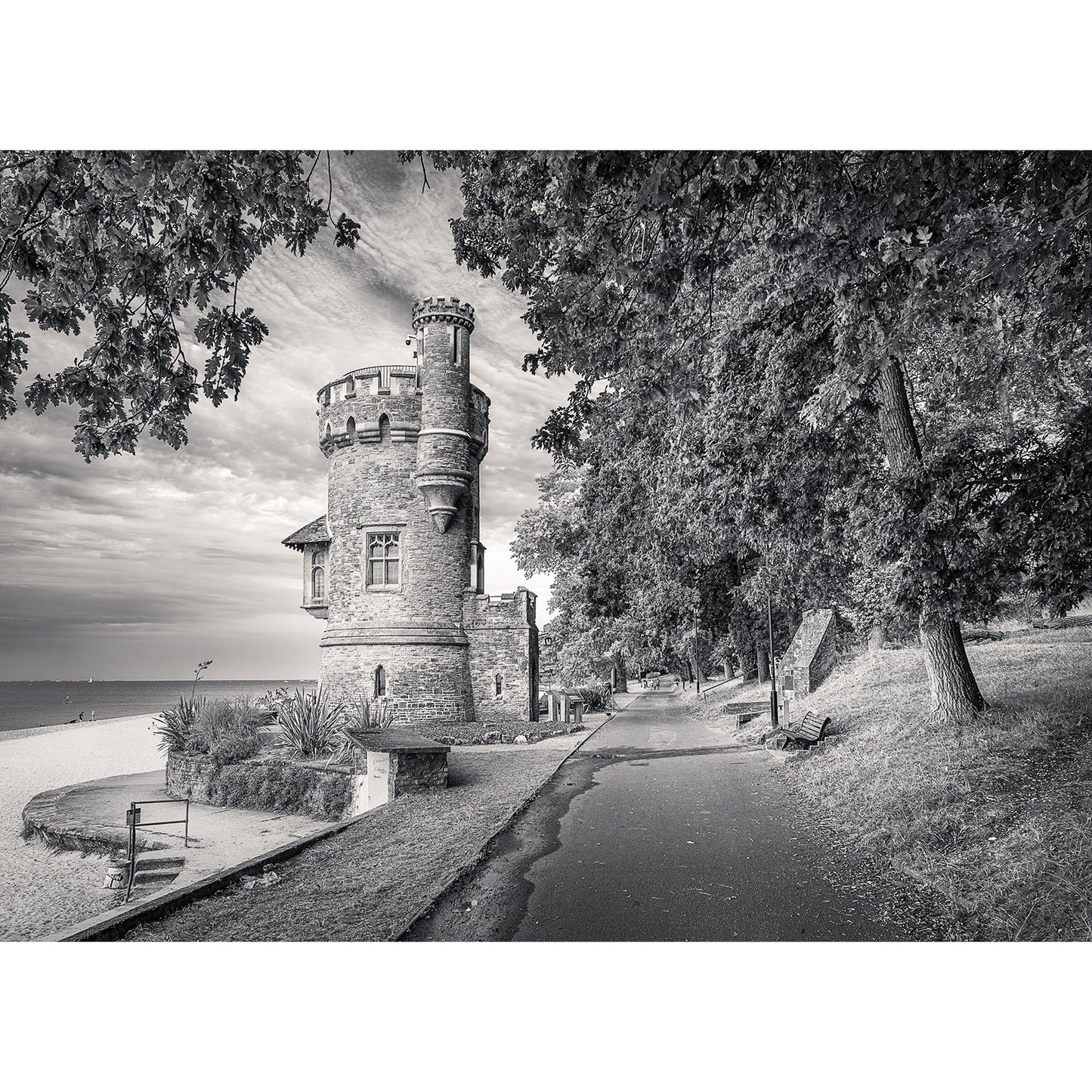 A historic stone tower named Appley Tower stands by the edge of a tree-lined path leading to the beach, with the sea visible in the background under a cloudy sky. Image number 2780 by Available Light Photography captures this timeless scene beautifully.