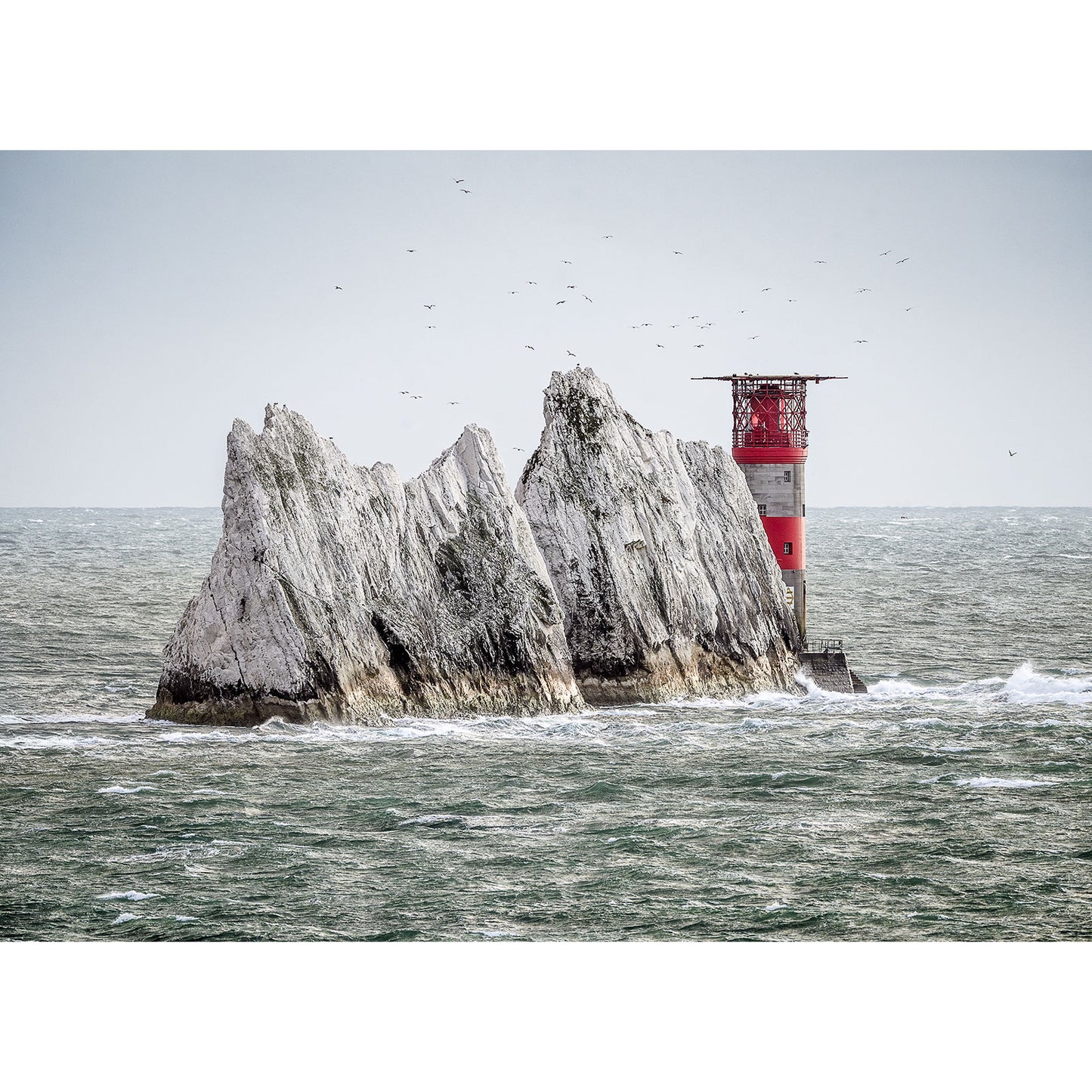 A scenic view of The Needles by Available Light Photography captures a picturesque red and white lighthouse perched on a rocky outcrop in the sea, with waves crashing against the rocks and seabirds soaring above, creating ideal conditions for photography.