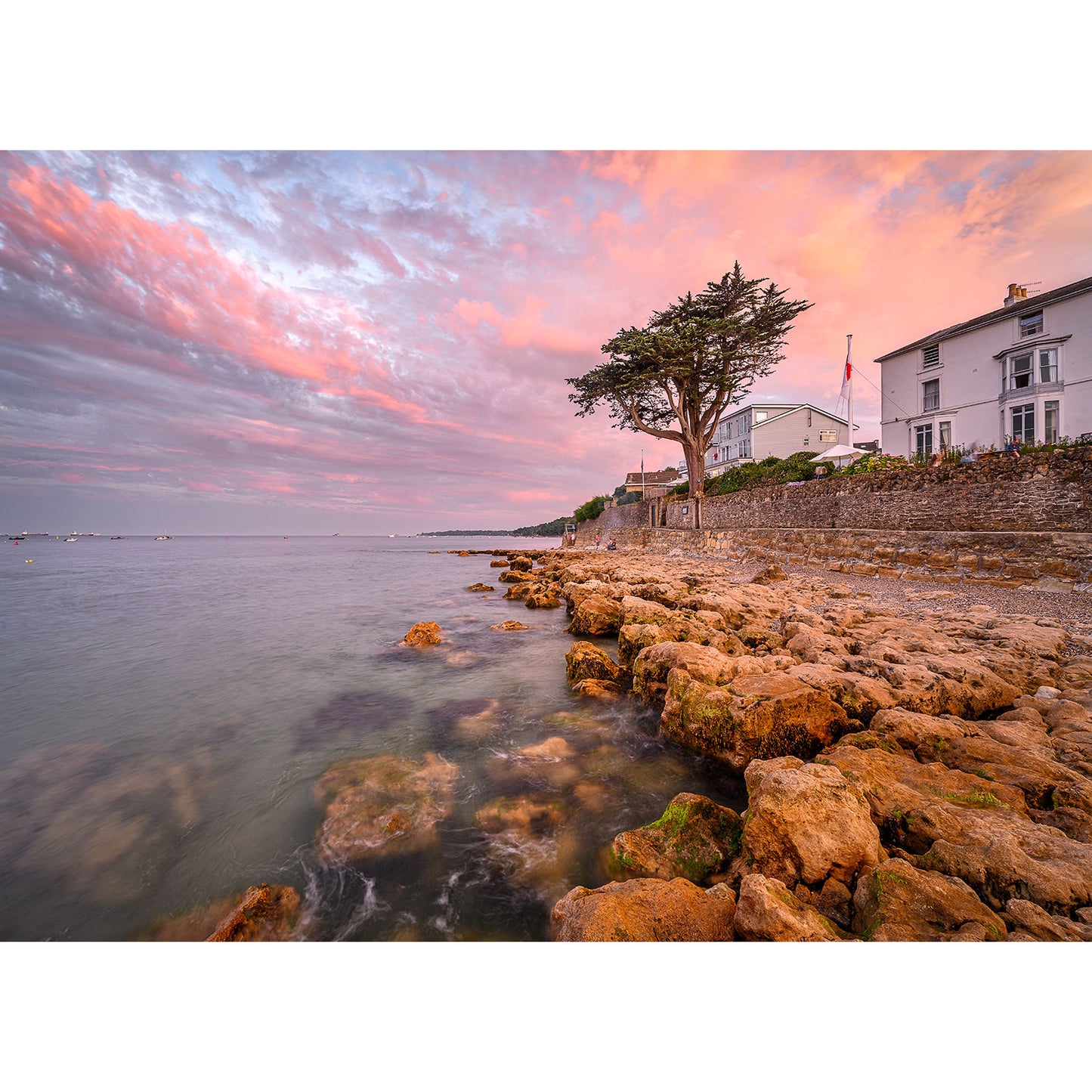 Experience the serenity of a coastal sunset with "Seaview" by Available Light Photography, featuring a rocky shore in the foreground, a calm sea to the left, and charming houses along a stone wall to the right under a vibrant sky. This idyllic scene truly embodies nature's tranquility as captured in image number 1196.