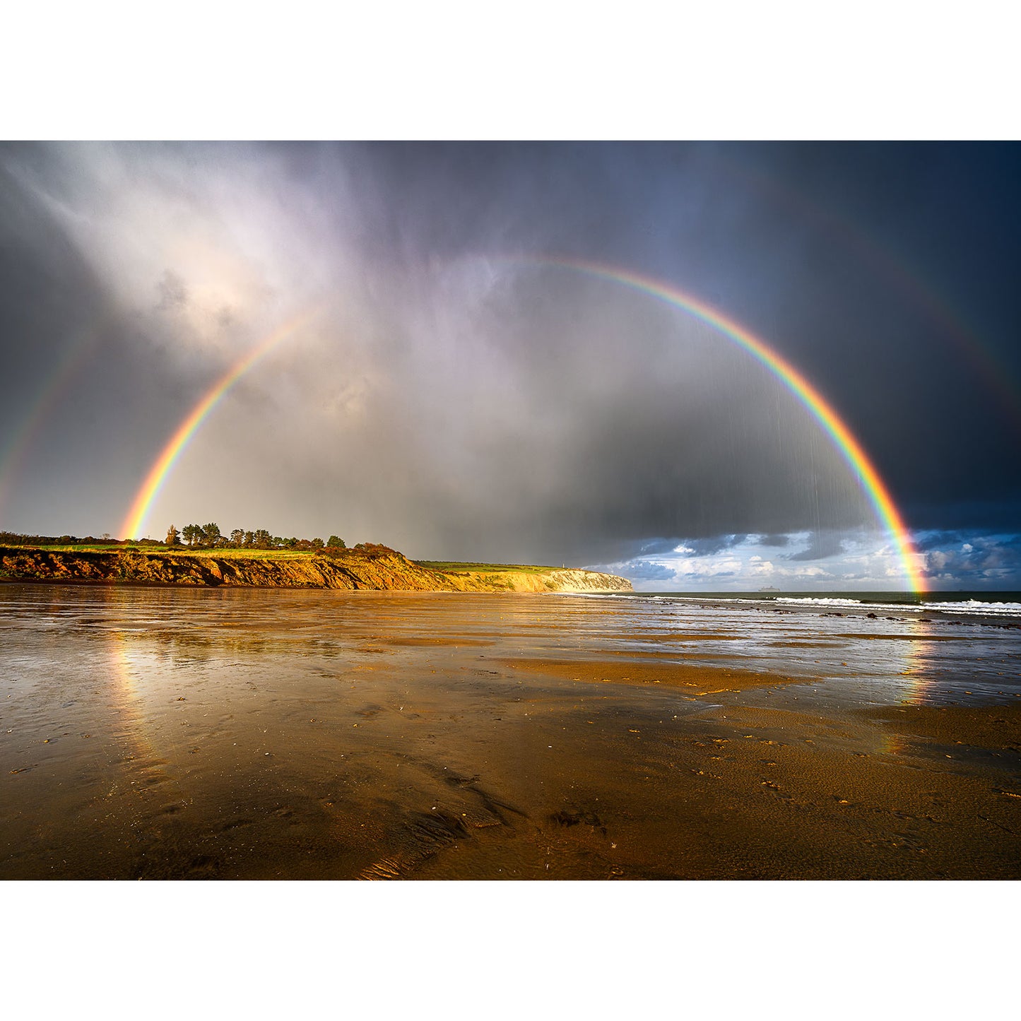A stunning photograph titled "Rainbow over Culver Cliff" by Available Light Photography captures a double rainbow beautifully arching over a sandy beach, as dark clouds recede, revealing the coastline and ocean in the background.