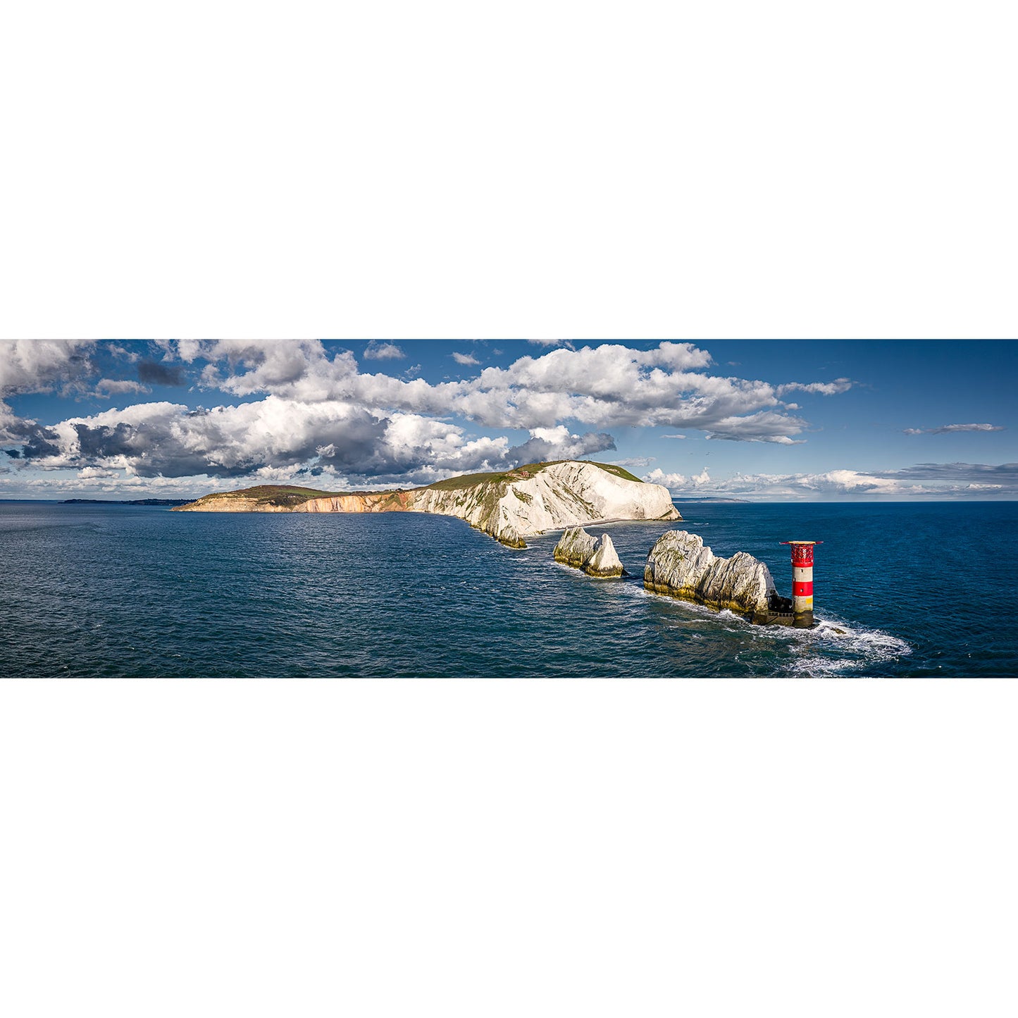 A photograph by Available Light Photography captures a panoramic view of The Needles’ rocky shoreline, highlighting the famous white cliffs and the red and white lighthouse on the right, set against a backdrop of blue sea and partly cloudy sky.
