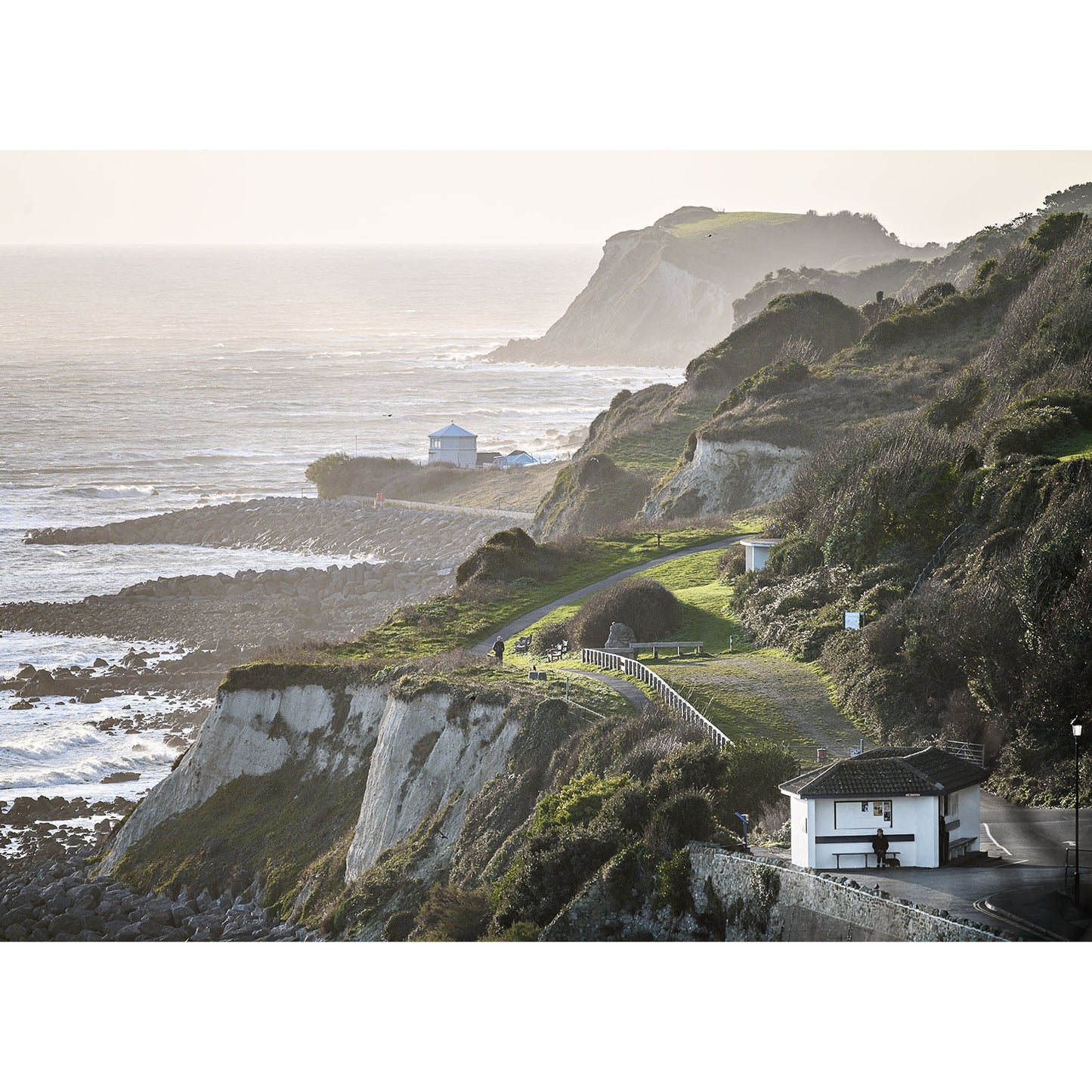 The breathtaking photograph "Looking toward Steephill Cove" by Available Light Photography captures a stunning coastal landscape. It features majestic cliffs overlooking the ocean, complemented by a charming building in the foreground and serene rolling hills gently unfurling under a hazy sky. Perfect for those seeking coastal beauty and nature's tranquility, this piece beautifully merges elements of nature and architecture.