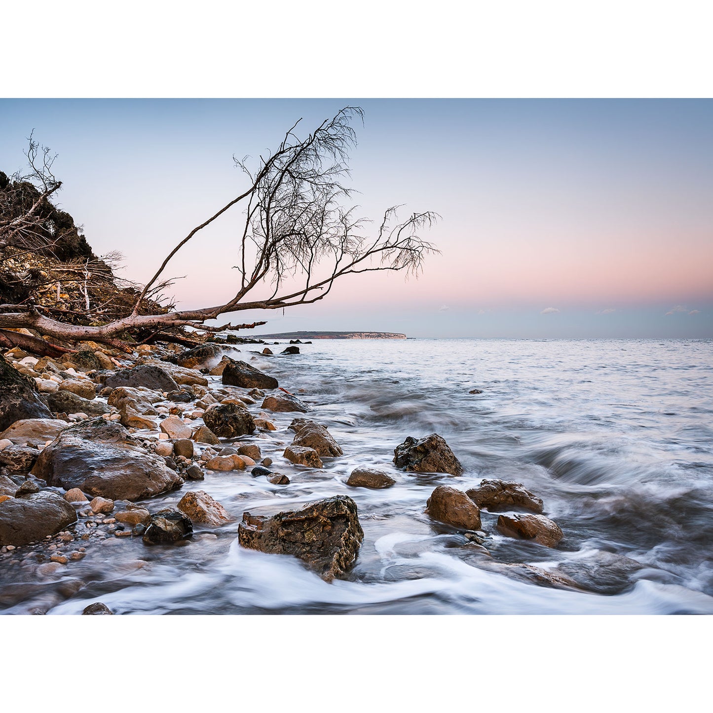 Steel Bay by Available Light Photography captures a rocky shoreline adorned with driftwood, where gentle waves kiss the coast at sunset, creating a serene and picturesque scene that evokes tranquility.