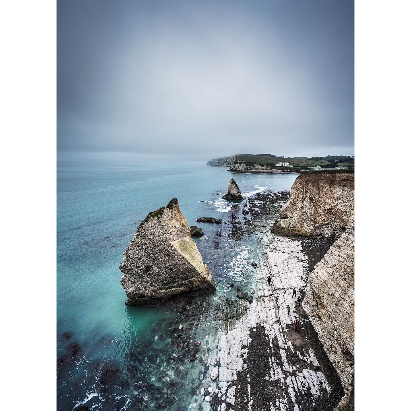 The breathtaking vantage point of Freshwater Bay by Available Light Photography captures rocky cliffs and majestic sea stacks rising from the ocean under a cloudy sky, waves gently lapping the rugged shoreline.