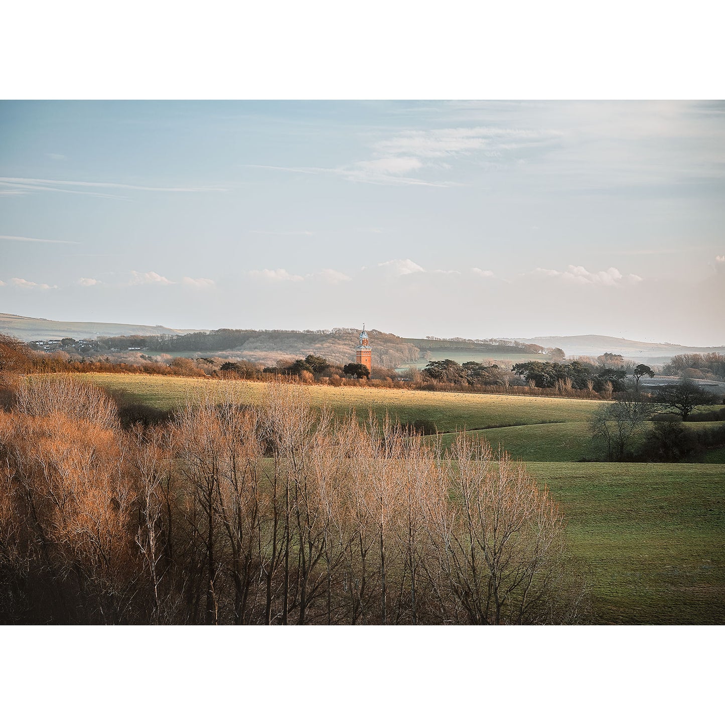 Whitecroft, Gatcombe by Available Light Photography captures a tranquil rural scene of a tall church tower amid rolling fields and trees beneath a partly cloudy sky.
