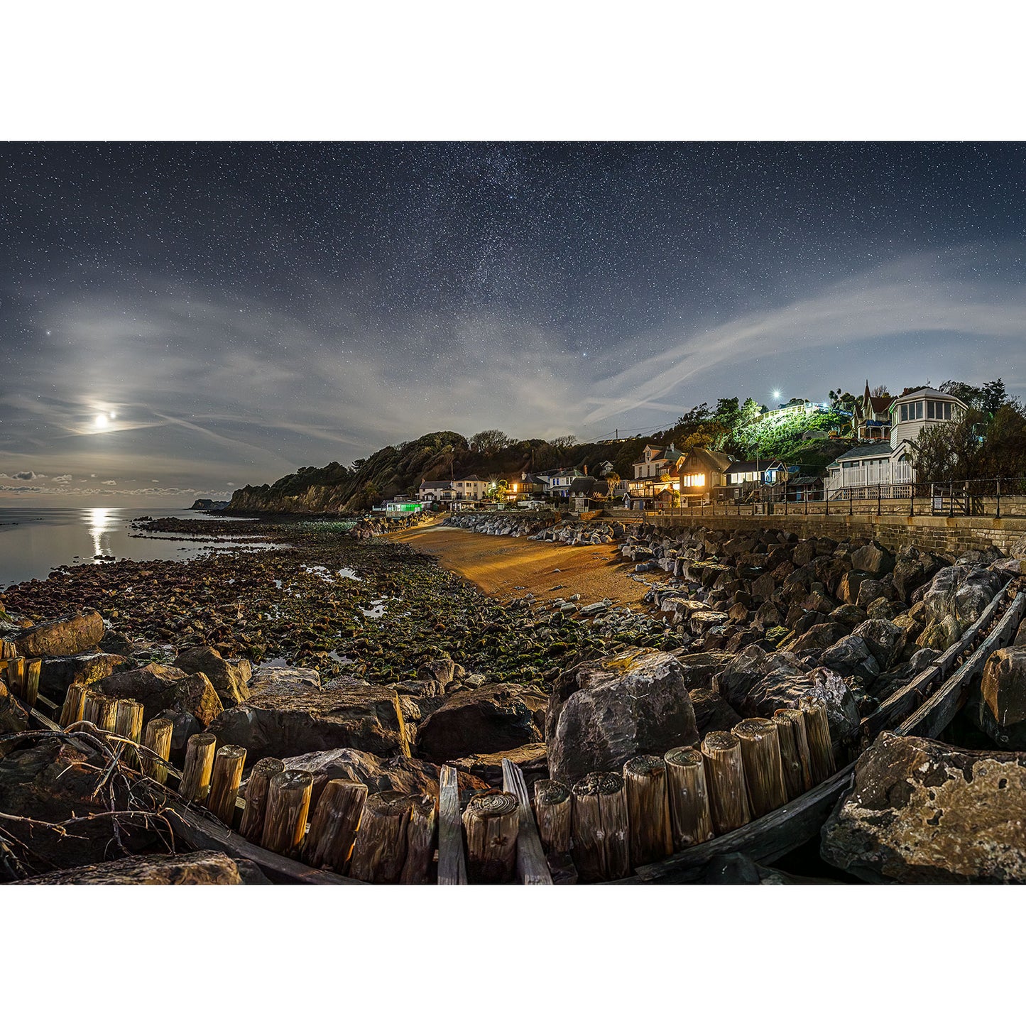 The "Steephill Cove" by Available Light Photography captures a serene coastal night with rocky shores and charming, glowing hillside houses. Moonlight and stars illuminate the sky, casting reflections on tranquil waters, creating a picturesque atmosphere.