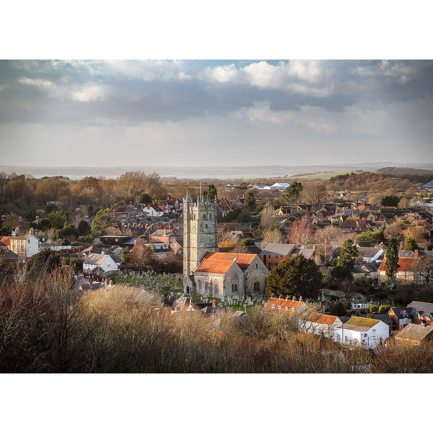 Under a cloudy sky, Carisbrooke by Available Light Photography captures an aerial view of a town. A prominent stone church with a tall tower stands surrounded by houses and trees, creating a picturesque scene in image number 2698.