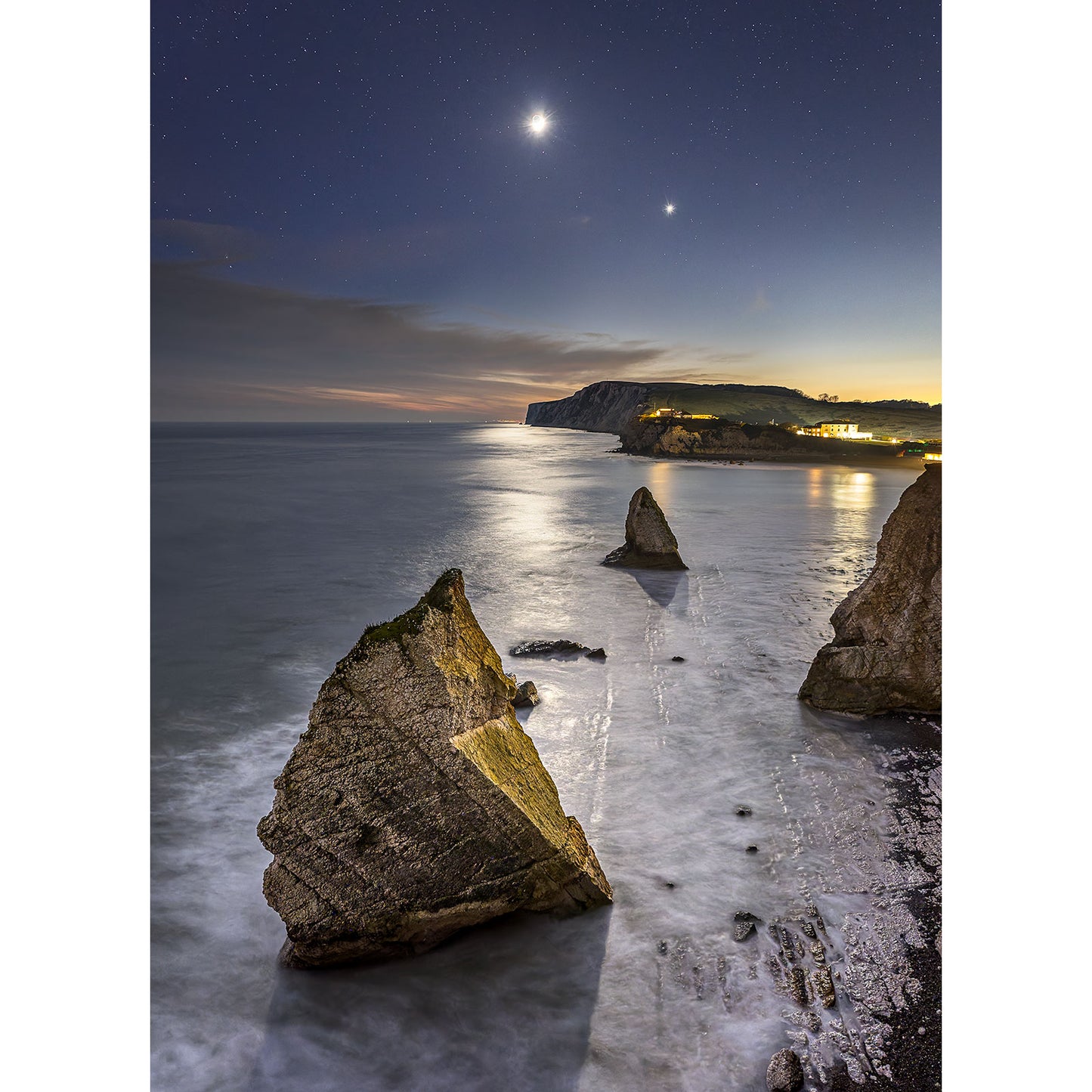 In "Freshwater Bay" by Available Light Photography, the rocky coastline glimmers under a starlit sky. A crescent moon casts its serene glow over the calm sea, highlighting two prominent rocks in the foreground and softly illuminating distant cliffs for a tranquil, mesmerizing scene.