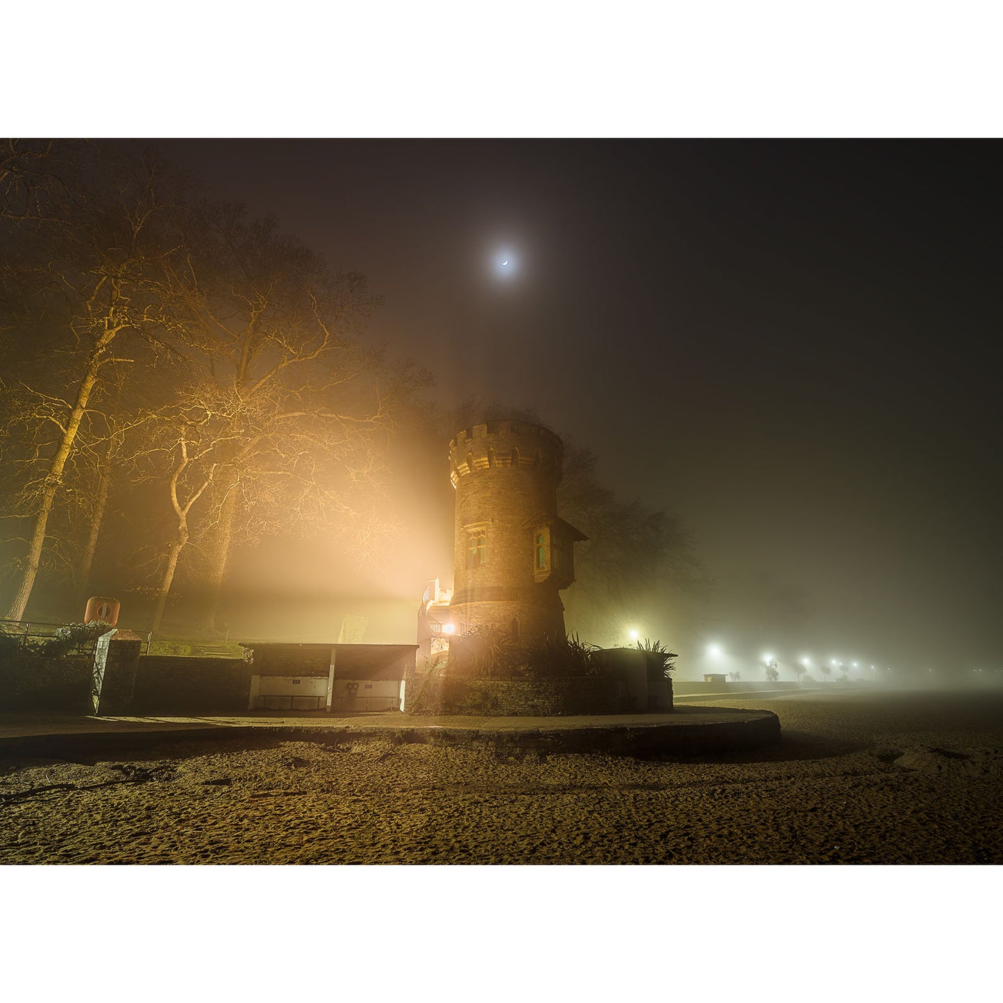 In a foggy landscape under a faint moon, Appley Tower from Available Light Photography stands amidst bare trees on sandy ground, its warm lights glowing gently and creating a serene, mysterious atmosphere that makes one regret leaving it behind.