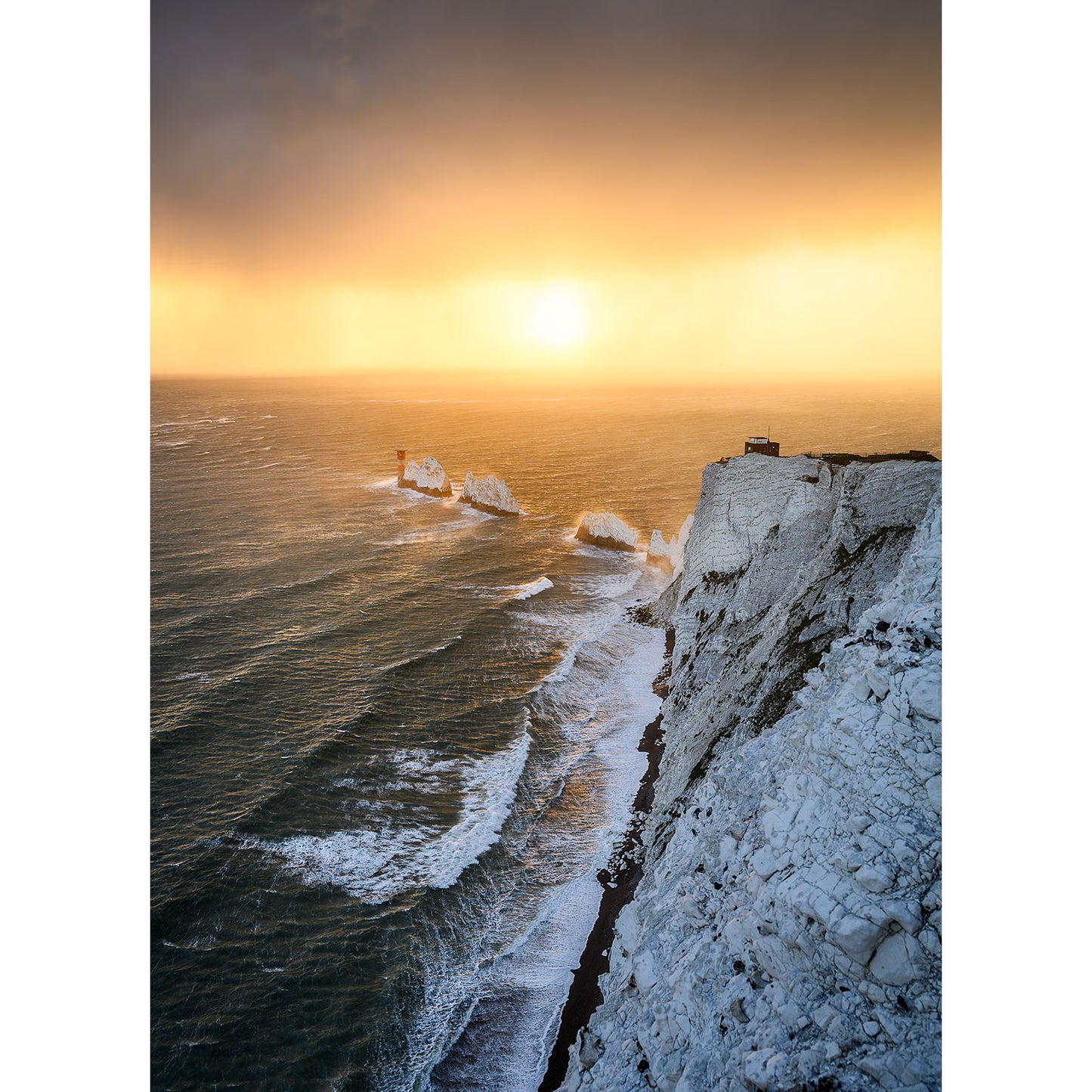 At sunset, The Needles’ white chalk cliffs tower over rough ocean waves, with sea stacks and a lighthouse beneath an orange sky. Captured by Available Light Photography.