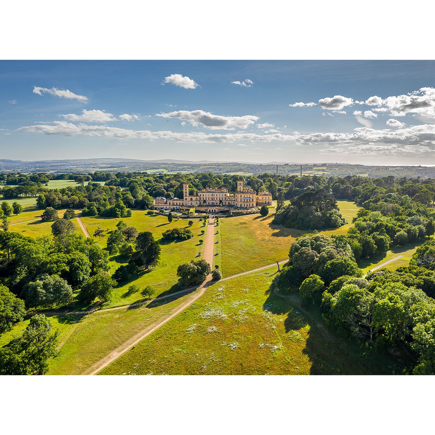 Aerial view of Osborne House by Available Light Photography captures Queen Victoria's Isle of Wight residence amid green lawns, trees, and winding paths beneath a blue, cloud-dotted sky.
