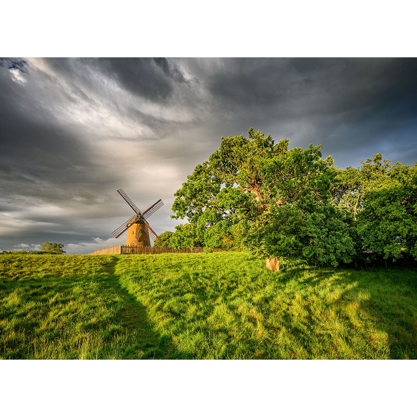 Available Light Photography captures Bembridge Windmill on a grassy hill beside lush trees under a cloudy sky, sunlight casting shadows across the field in a stunning drone shot.