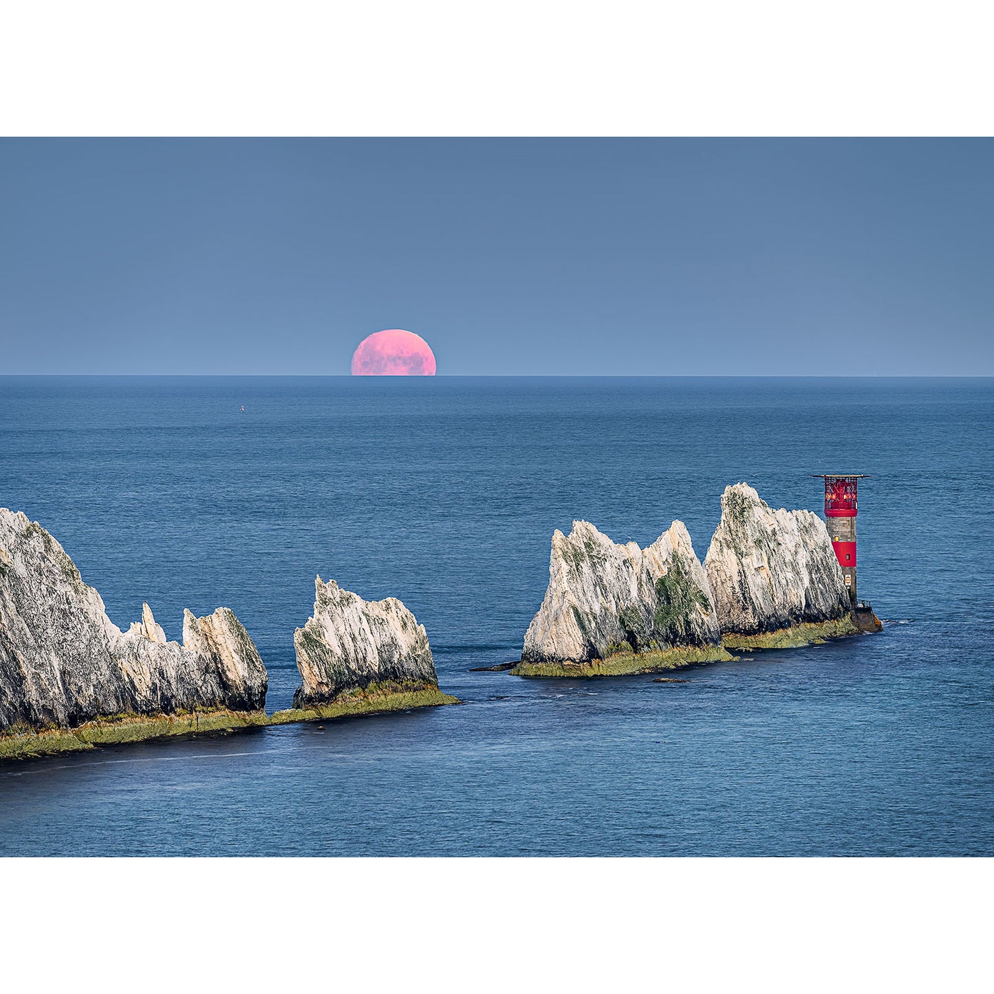 Moonset over The Needles by Available Light Photography captures jagged chalk rocks, a red and white lighthouse, and a pink full moon rising above the blue sea in a stunning seascape.