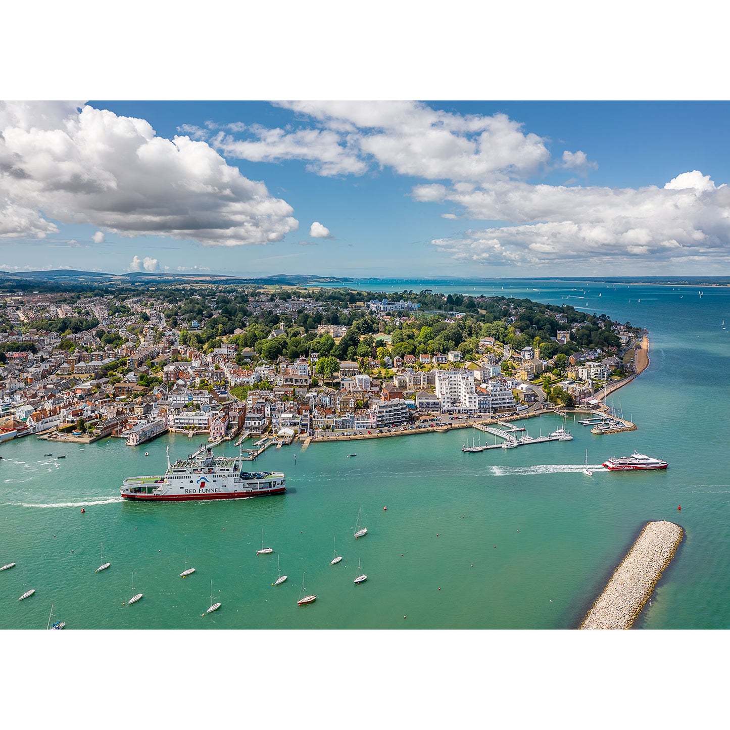 Aerial view of Cowes by Available Light Photography shows boats and ferries in the water, waterfront buildings, and green hills under a partly cloudy sky—perfect wall art for coastal or nautical decor.