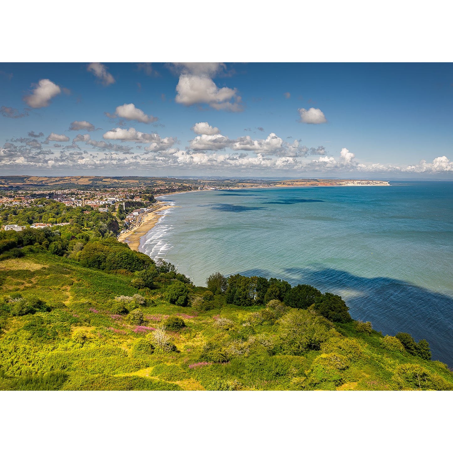 Sandown Bay from Luccombe by Available Light Photography features green hills, a sandy shoreline, and a coastal town under a blue sky with clouds.