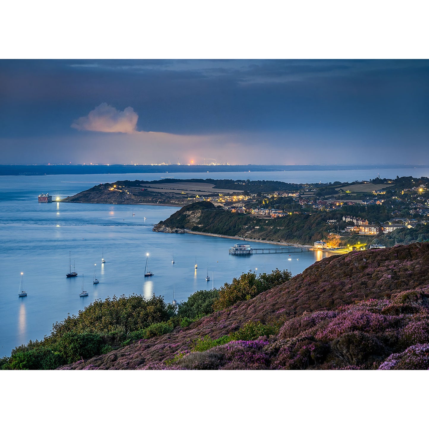 Totland by Available Light Photography captures a coastal landscape at dusk with illuminated houses along the shore, anchored boats in calm water, and distant city lights on the horizon.