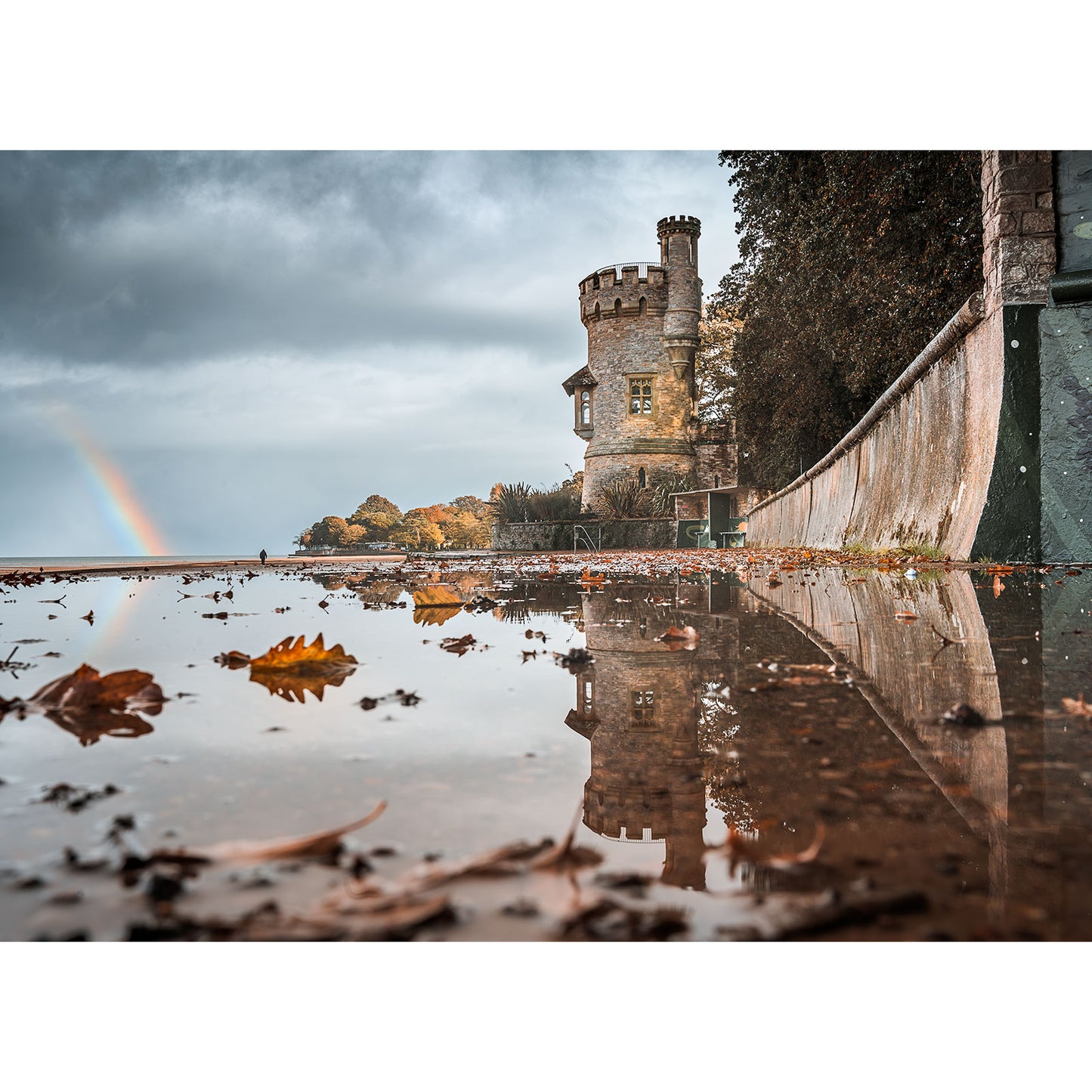 Autumn, Appley Tower by Available Light Photography captures a stone tower and wall reflected in a large puddle, autumn leaves scattered around, and a faint rainbow visible in the cloudy sky.