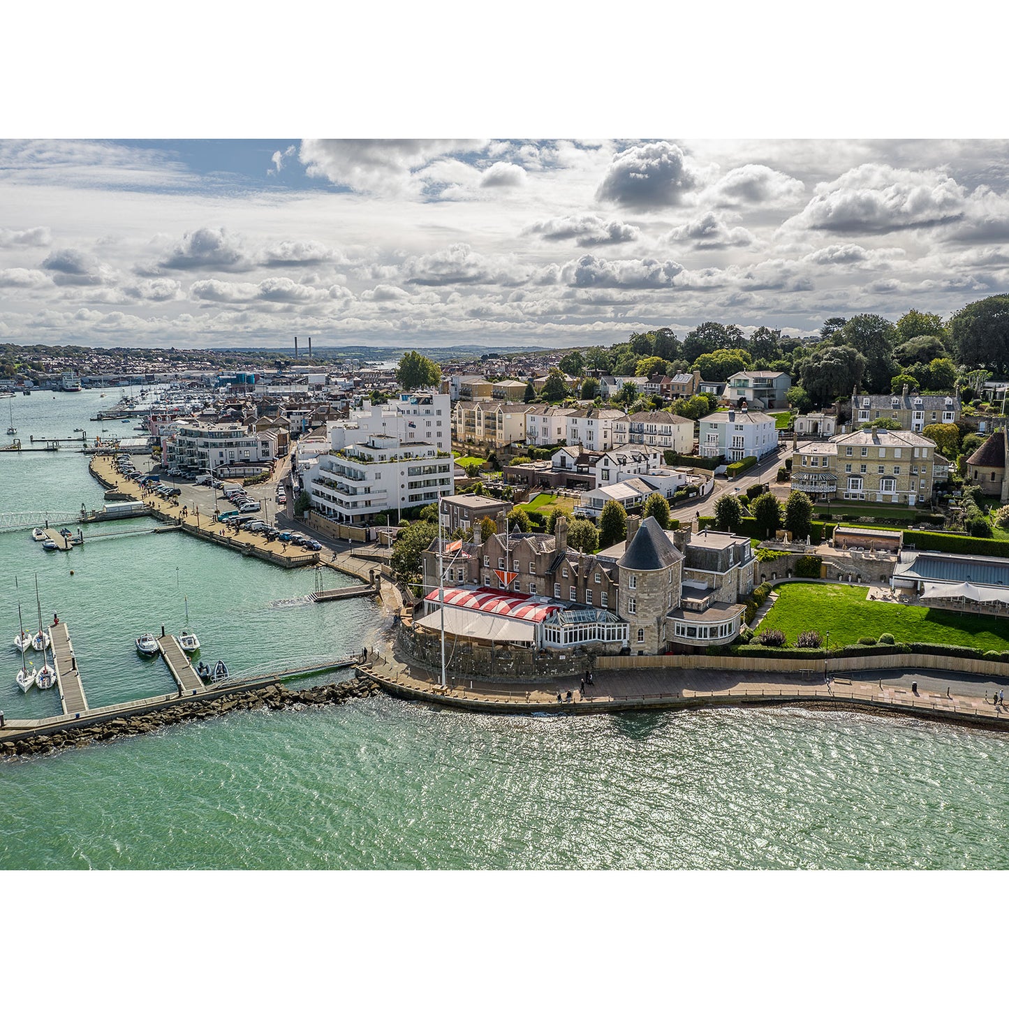 Aerial view of The Royal Yacht Squadron, Cowes by Available Light Photography shows vibrant marinas, waterfront buildings, and sleek boats docked along the shoreline beneath a partly cloudy sky.