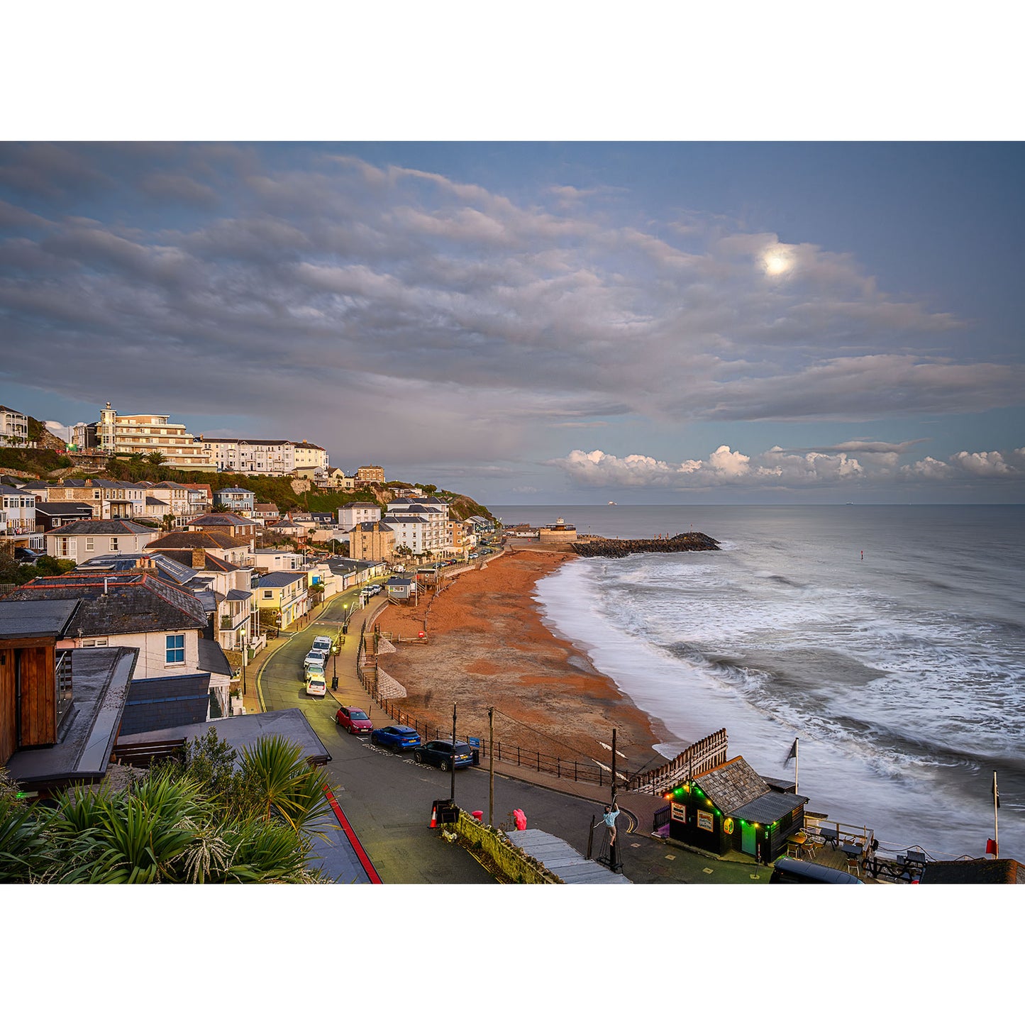 Moonrise over Ventnor by Available Light Photography shows a coastal town with buildings along a curved road beside a sandy beach and calm sea under a cloudy sky at sunset, as seen in image 3100.