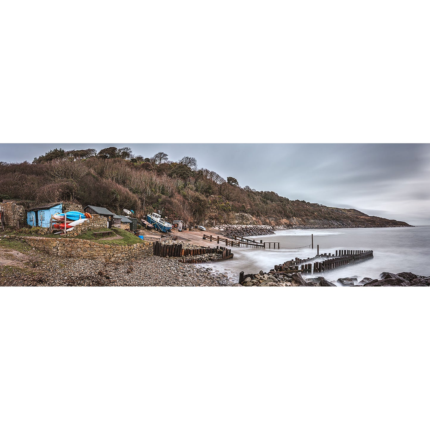 Castlehaven by Available Light Photography captures a panoramic rocky shoreline with boats, wooden groynes, charming buildings, and a tree-covered hill under an overcast sky.