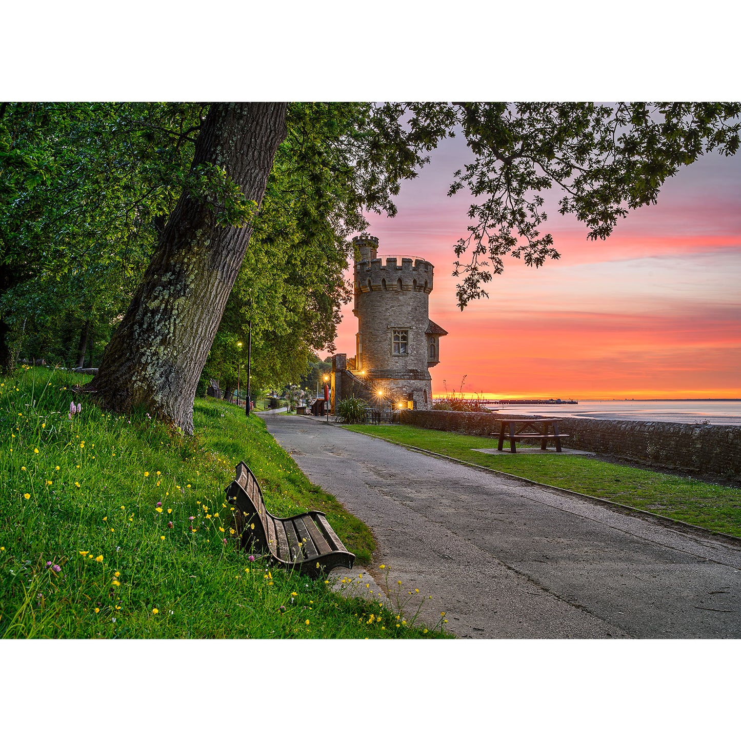 Appley Tower by Available Light Photography captures a stone tower beside a serene waterfront at sunset, with a tree-lined path, benches, and grassy foreground dotted with yellow flowers.