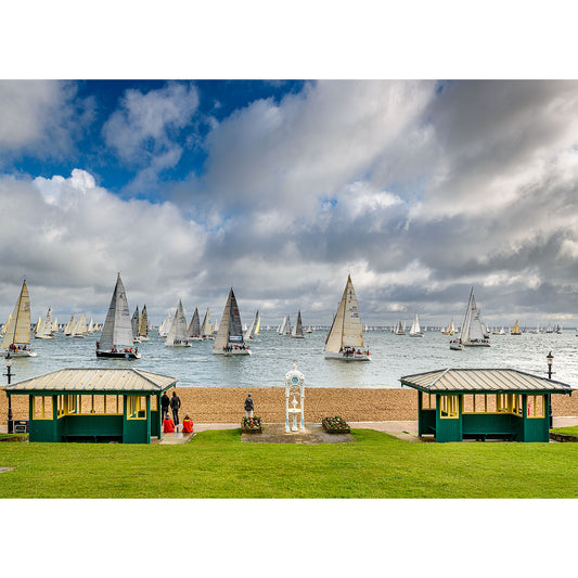 Sailboats participating in the Round the Island Race regatta under a dynamic cloudy sky, viewed from the Isle of Wight's shore with green huts, captured by Available Light Photography.