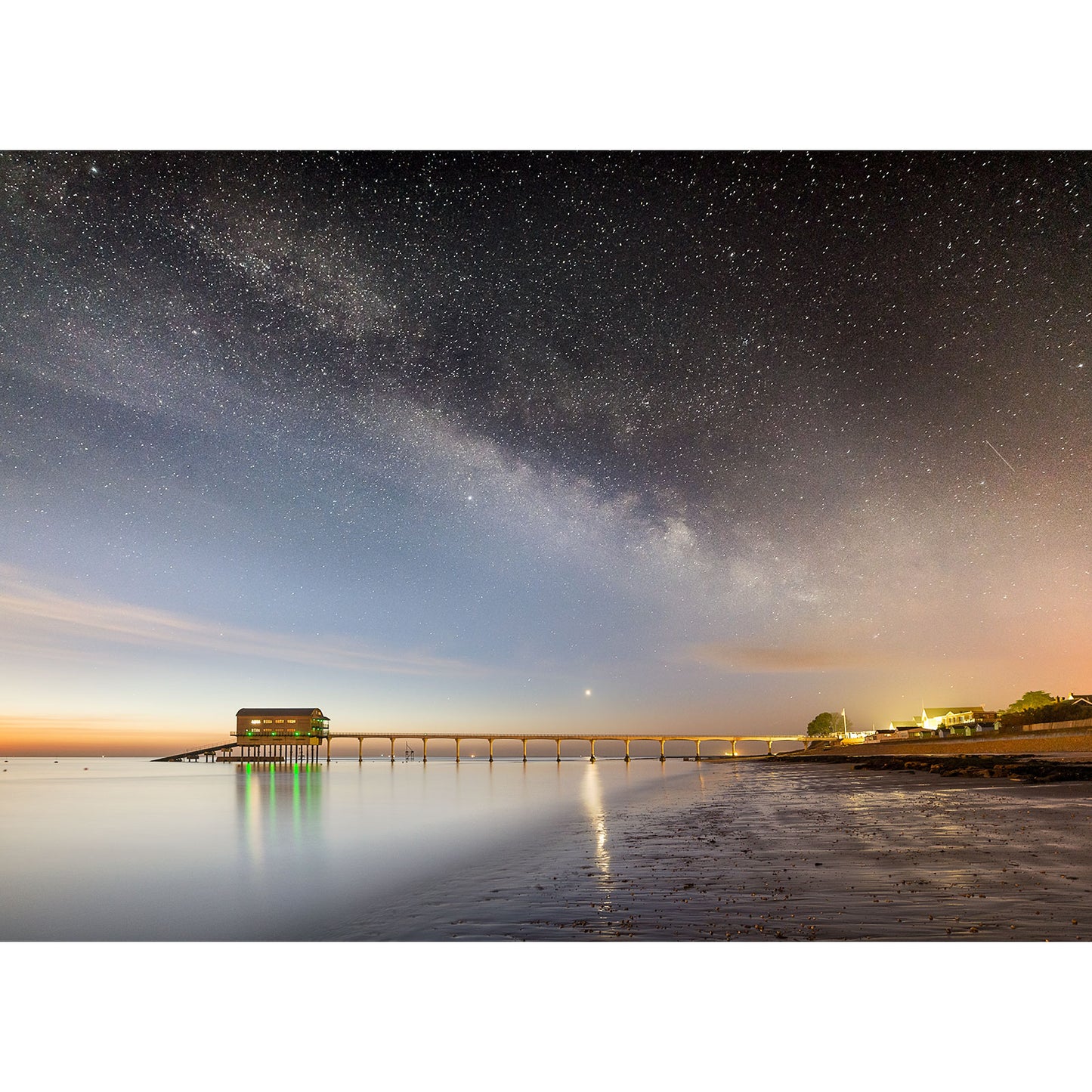 A Milky Way night sky over the serene Isle of Wight beach with a long pier illuminated by lights reflecting on the water's surface captured by Available Light Photography at Bembridge Lifeboat Station.