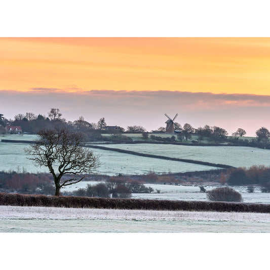 A frost-covered landscape at dawn with Bembridge Windmill on the horizon under a gradient orange sky on the Isle of Wight.