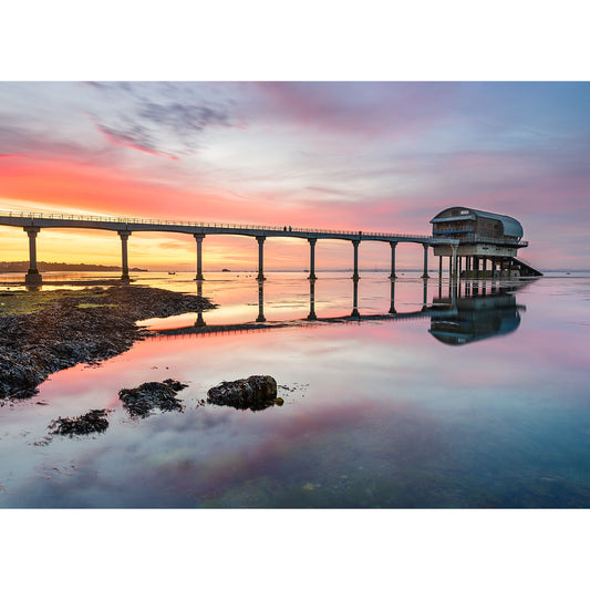 A picture of the Bembridge Lifeboat Station with a pier leading to an overwater building against a vibrant sunset sky, reflected on the Isle of Wight's water's surface by Available Light Photography.