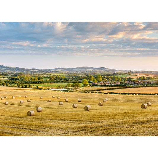 A tranquil rural landscape featuring an Arreton Valley field dotted with hay bales under a soft sky at sunset, with a view of a village and rolling hills in the distance, captured by Available Light Photography.