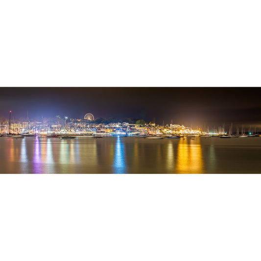 A panoramic nighttime cityscape of Cowes, a coastal city on the Isle of Wight, with illuminated buildings and reflections on the water by Available Light Photography.