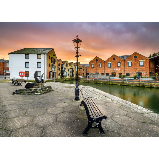 Sunset over a tranquil harbor with historic warehouse buildings, a cobblestone promenade, a wooden bench, and a classic street lamp on the Isle of Wight captured by Newport Quay from Available Light Photography.