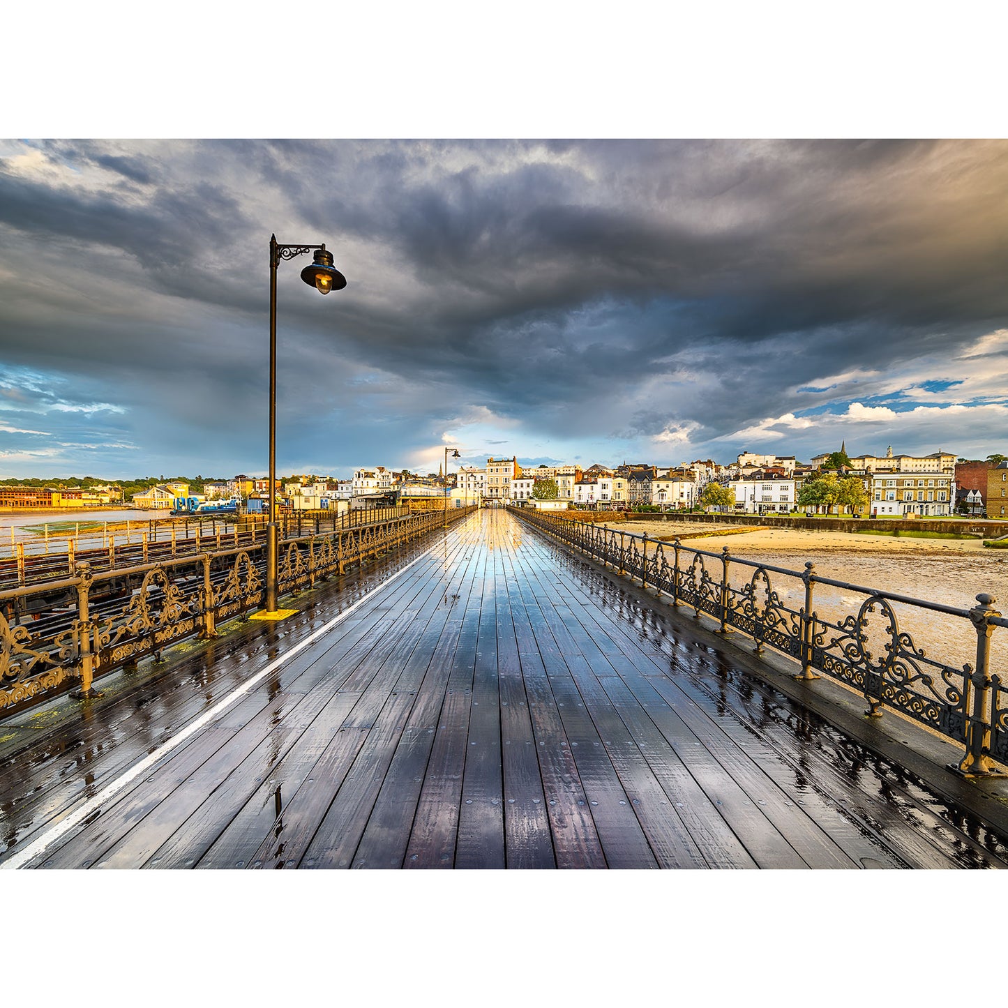 A wet Ryde Pier leading to a coastal town on the Isle of Wight under a dramatic sky at sunset.