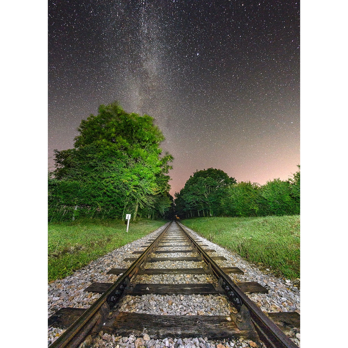 Railroad tracks leading into a forest under a starry night sky, reminiscent of a Milky Way designed by Steve Gascoigne at Ashey Junction by Available Light Photography.